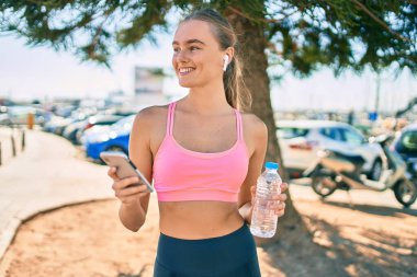 Genç sarışın bayan akıllı telefon kullanıyor ve şehirde bir şişe su içiyor..