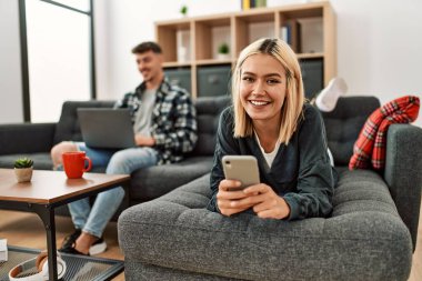 Evdeki kanepede oturan genç beyaz çift dizüstü bilgisayar ve akıllı telefon kullanarak mutlu bir şekilde gülümsüyor..