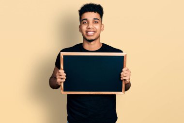 Afro-Amerikalı genç adam elinde karatahtayla mutlu bir şekilde ayakta duruyor ve gülümsüyor. 