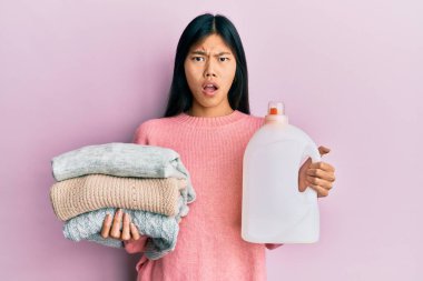 Genç Çinli bir kadın elinde deterjan şişesi ve şok olmuş giysilerle kuşkucu ve alaycı görünüyor. 