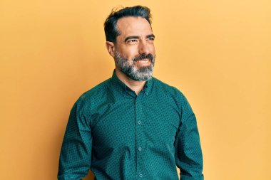 Sakallı, gri saçlı, iş kıyafetleri giyen, yüzünde gülümsemeyle yan yana bakan orta yaşlı bir adam. Kendinden emin gülen. 