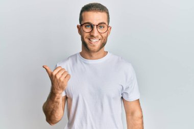 Gündelik kıyafetler ve gözlük takan genç beyaz bir adam gülümseyen bir yüzle yan tarafa bakıyor ve başparmağıyla işaret ediyor.. 