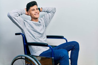 Tekerlekli sandalyede oturan genç adam rahatlıyor ve esniyor, kolları ve elleri başının arkasında mutlu bir şekilde gülümsüyor. 