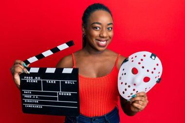 Genç Afro-Amerikan kadın elinde video film panosu ve korku maskesi ile gülümseyen mutlu ve havalı bir gülümsemeyle. diş gösterme. 