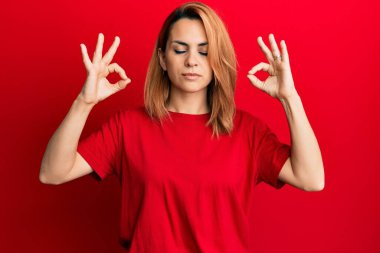 Latin kökenli genç bir kadın, gündelik kırmızı tişört giyip rahatlıyor ve gözleri kapalı gülümsüyor parmaklarıyla meditasyon hareketi yapıyor. yoga konsepti. 