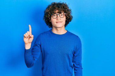 Yakışıklı genç adam günlük kıyafetler ve gözlük takıyor. Bir numarayı gösteriyor ve işaret ediyor. Kendine güvenen ve mutlu bir şekilde.. 