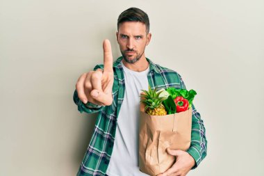 Sakallı yakışıklı bir adam elinde poşetle bakkaliyeyi işaret ediyor ve kızgın bir ifade takınıyor. 