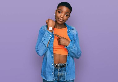 Genç Afro-Amerikan kadın günlük kıyafetler giyiyor. Aceleyle saati, sabırsızlığı ve rahat bir ifadeyle kameraya bakıyor. 