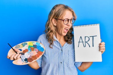 Orta yaşlı sarışın kadın boyacı paleti ve defter tutuyor kızgın ve kızgın bağırıyor öfkeyle bağırıyor. öfke ve agresif konsept. 