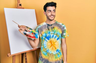 Genç İspanyol adam paletle resim çiziyor, resim sehpası ayakta gülümsüyor, yan tarafa bakıyor ve düşünüyor.. 