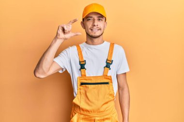 Tamirci üniforması giyen, gülümseyen, kendine güvenen, el hareketi yapan, parmaklarıyla işaret yapan, kameraya bakan İspanyol bir genç. ölçüm kavramı. 