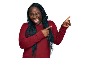 Saçları örgülü genç siyahi bir kadın gülümseyerek kameraya bakıyor ve iki eliyle ve parmaklarıyla kenarı işaret ediyor.. 