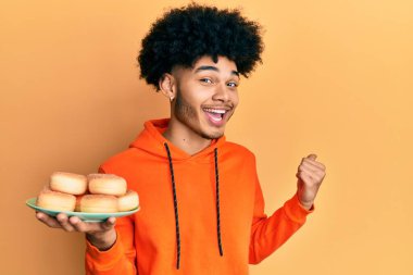 Afro saçlı genç Afrikalı Amerikalı adam elinde lezzetli şekerli çöreklerle başparmağını yana uzatarak mutlu bir şekilde gülümsüyor. 