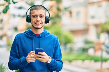 Genç İspanyol adam şehirde akıllı telefon ve kulaklık kullanarak mutlu gülümsüyor..
