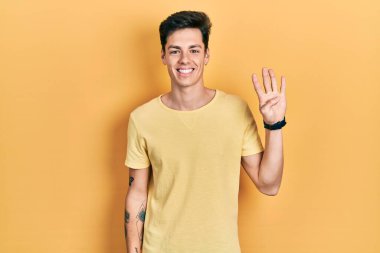 İspanyol asıllı genç adam sarı bir tişört giyiyor. Dört numarayı gösteriyor ve işaret ediyor. Kendinden emin ve mutlu bir şekilde gülümsüyor.. 