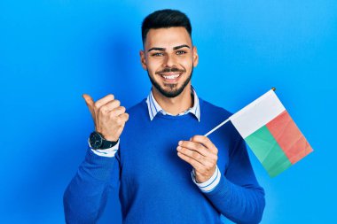 Sakallı genç İspanyol bir adam elinde madagaskar bayrağı başparmağıyla yan tarafa bakıyor ve ağzı açık gülümsüyor. 