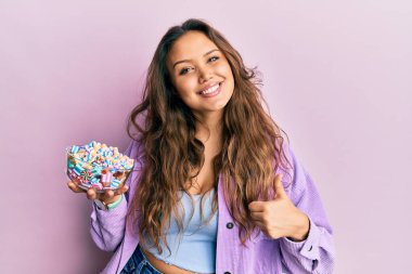 Genç İspanyol kız elinde şeker dolu bir kaseyle mutlu ve pozitif gülümsüyor, başparmağıyla mükemmel ve onay işareti yapıyor. 