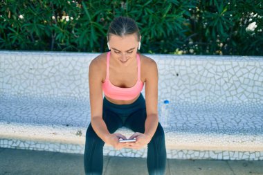 Genç sarışın sporcu, parkta bankta oturan akıllı telefonu kullanarak egzersiz yapıyor..