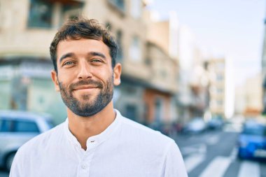 Güneşli bir günde günlük beyaz gömlek giyen sakallı yakışıklı adam dışarıda mutlu bir şekilde gülümsüyor.