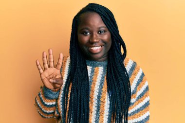 Saçları örgülü genç siyahi bir kadın günlük kışlık süveter giyiyor ve parmaklarıyla dört numarayı gösterirken kendinden emin ve mutlu gülümsüyor.. 