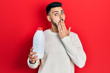 Sakallı, elinde litrelik süt şişesi olan, ağzı elle kaplanmış, şok olmuş ve hata yapmaktan korkan genç bir İspanyol. şaşırmış ifade 