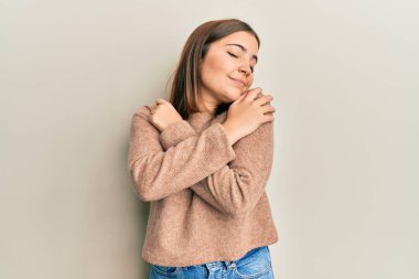 Genç ve güzel bir kadın gündelik kıyafetler giyip kendine sarılıyor mutlu ve pozitif, kendinden emin gülümsüyor. Sevgi ve özgüven. 