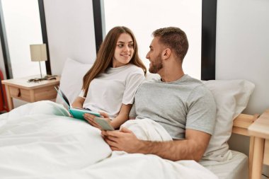 Genç beyaz çift kitap okuyor ve yatakta akıllı telefon kullanıyor..
