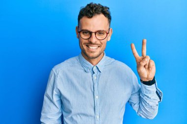 Gündelik kıyafetler giyen ve gözlüklü genç İspanyol adam iki numarayı gösterip işaret ederken kendinden emin ve mutlu bir şekilde gülümsüyor.. 