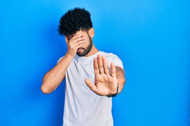 Sakallı, günlük beyaz tişörtlü, elleriyle gözlerini kapatan üzgün ve korku ifadeli genç bir Arap adam. utangaç ve olumsuz kavram. 