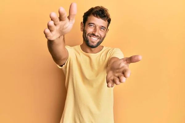 Handsome man with beard wearing casual yellow tshirt over yellow background looking at the camera smiling with open arms for hug. cheerful expression embracing happiness.