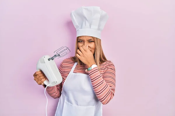 Beautiful hispanic woman holding pastry blender electric mixer smelling something stinky and disgusting, intolerable smell, holding breath with fingers on nose. bad smell
