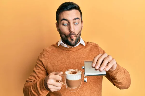 Young hispanic man drinking a cup of coffee with alcohol making fish face with mouth and squinting eyes, crazy and comical.