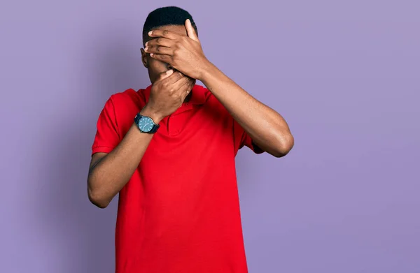 目と口を手で覆い 驚きと衝撃を受けたカジュアルな赤いTシャツを着た若いアフリカ系アメリカ人男性 感情を隠す — ストック写真
