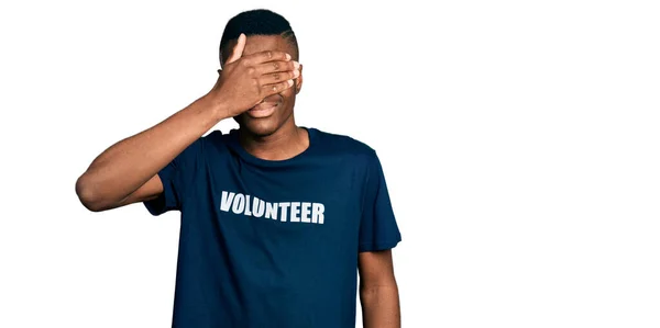 ボランティアのTシャツを着た若いアフリカ系アメリカ人の男性が笑顔で 驚きのために目を覆う顔を手で笑っています 視覚障害の概念 — ストック写真