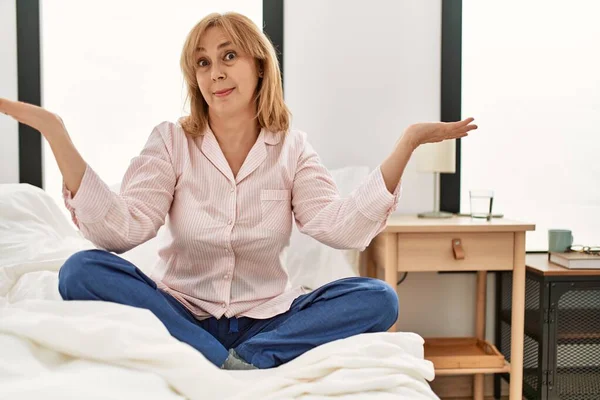 中年の白人女性はベッドの上に座っパジャマを着て 腕や手を上げて混乱した表情 疑わしい概念 — ストック写真
