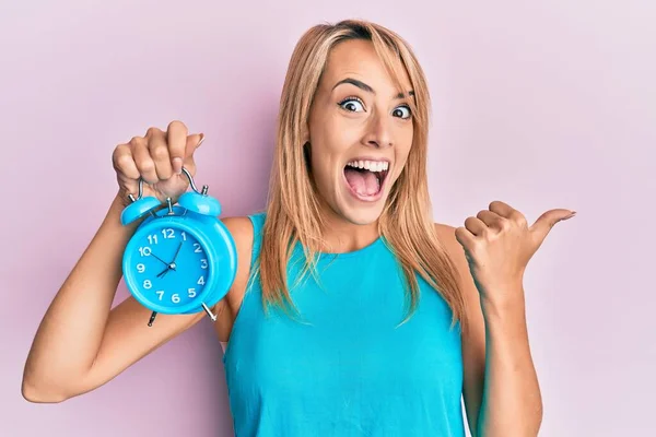 Beautiful Blonde Woman Holding Alarm Clock Pointing Thumb Side Smiling — ストック写真