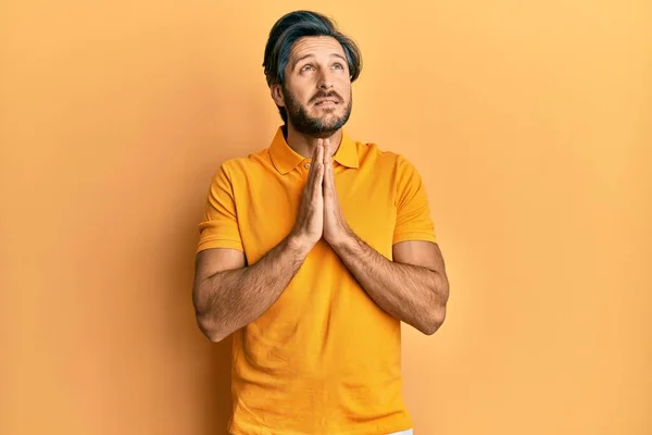 若いヒスパニック系の男性は カジュアルな黄色のTシャツを物乞いを着て手で祈ると一緒に顔に希望の表現は非常に感情的かつ心配している お願い — ストック写真