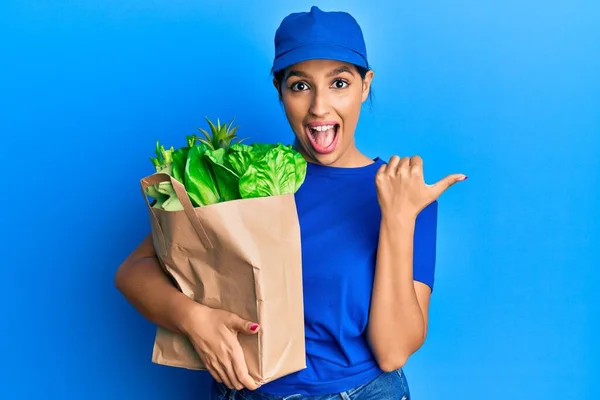 美しいですブルネット女性身に着けています宅配便の制服とともに食料品袋指して親指アップへザ側笑顔幸せとともにオープン口 — ストック写真