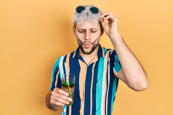 Young hispanic man with modern dyed hair drinking mojito glass making fish face with mouth and squinting eyes, crazy and comical.