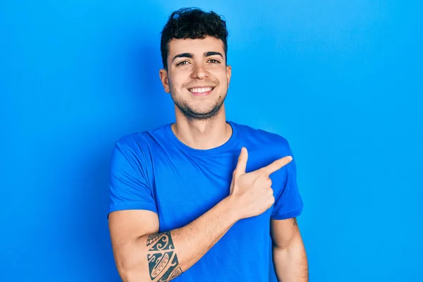 笑顔で陽気なカジュアルなブルーのTシャツを着た若いヒスパニック系の男が顔に幸せと自然な表情で側に手と指で指しています — ストック写真