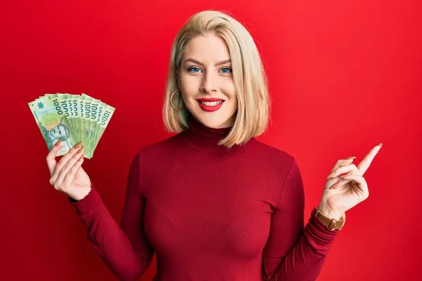 Young Blonde Woman Holding 1000 Chilean Pesos Smiling Happy Pointing — Stock Photo, Image