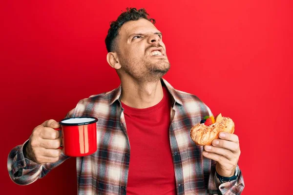 Young hispanic man drinking coffee and eating pastry angry and mad screaming frustrated and furious, shouting with anger. rage and aggressive concept.