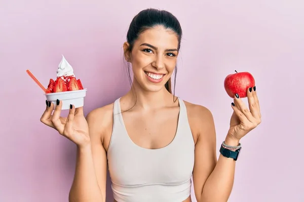 身穿运动服的年轻惊慌失措的女人 手里拿着苹果和冰淇淋 脸上挂着快乐而清凉的笑容 显示牙齿 — 图库照片