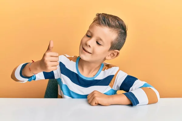 可敬的高加索小孩穿着休闲装坐在桌子上 一副骄傲自满的样子 微笑着向旁边竖起大拇指 — 图库照片