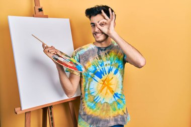 Genç İspanyol adam paletle resim çiziyor. Resim sehpası duruyor. El hareketleriyle gülümsüyor, gözleriyle mutlu yüzle parmaklarının arasından bakıyor.. 