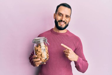 Sakallı genç adam çikolata parçacıklı kurabiye kavanozunu tutarken mutlu bir şekilde elini ve parmağını gösterdi. 