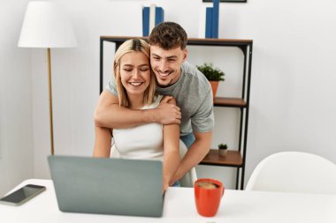 Genç, beyaz bir çift kucak açıp dizüstü bilgisayar kullanıyor. Evdeki masanın üstünde oturuyorlar..