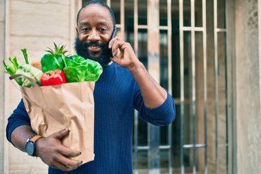 Elinde süpermarketten alınmış kağıt torba sakallı Afrikalı Amerikalı adam telefonda konuşuyor.