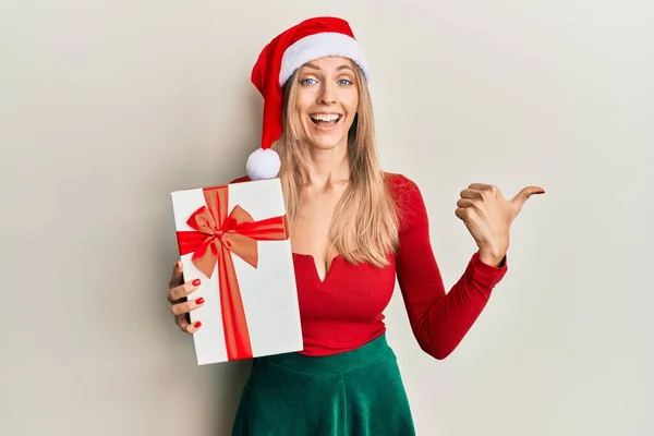 美丽的高加索女人头戴圣诞礼帽 手里拿着礼物 把大拇指指向旁边 笑容满面 张开嘴 — 图库照片