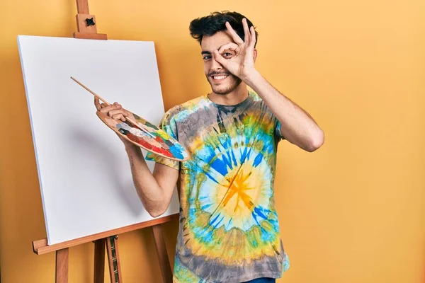 Genç İspanyol adam paletle resim çiziyor. Resim sehpası duruyor. El hareketleriyle gülümsüyor, gözleriyle mutlu yüzle parmaklarının arasından bakıyor.. 